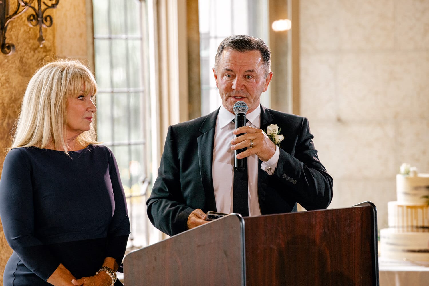 A man in a tuxedo speaks into a microphone while a woman listens beside him, set in a welcoming indoor venue with a cake in the background.