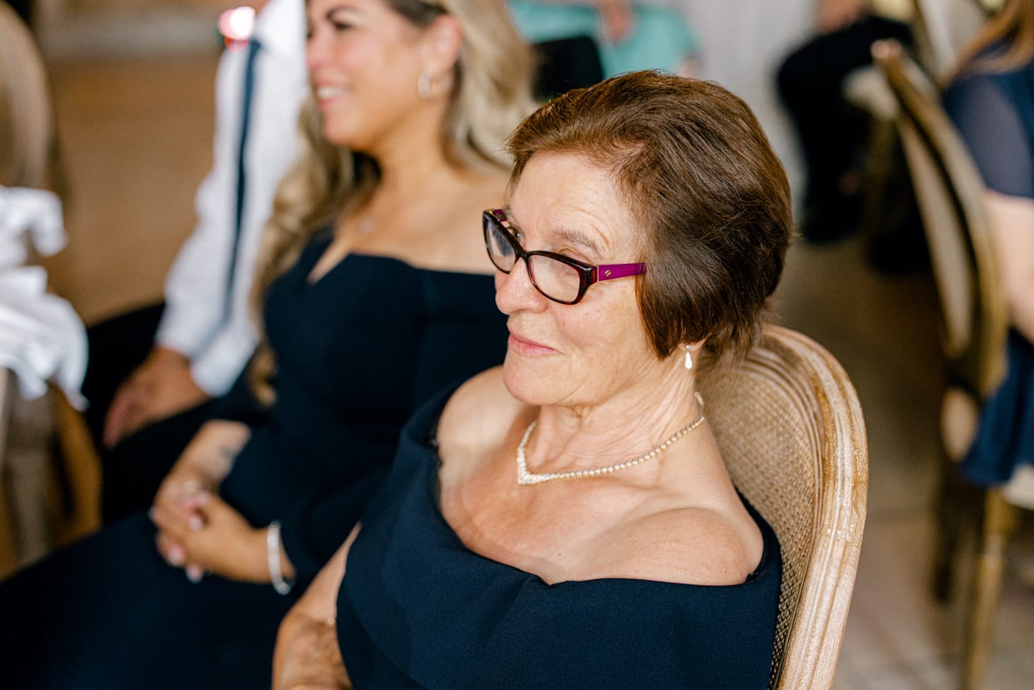 A woman in a black off-shoulder dress sits comfortably at a festive event, showcasing her elegant style with glasses and a delicate necklace.