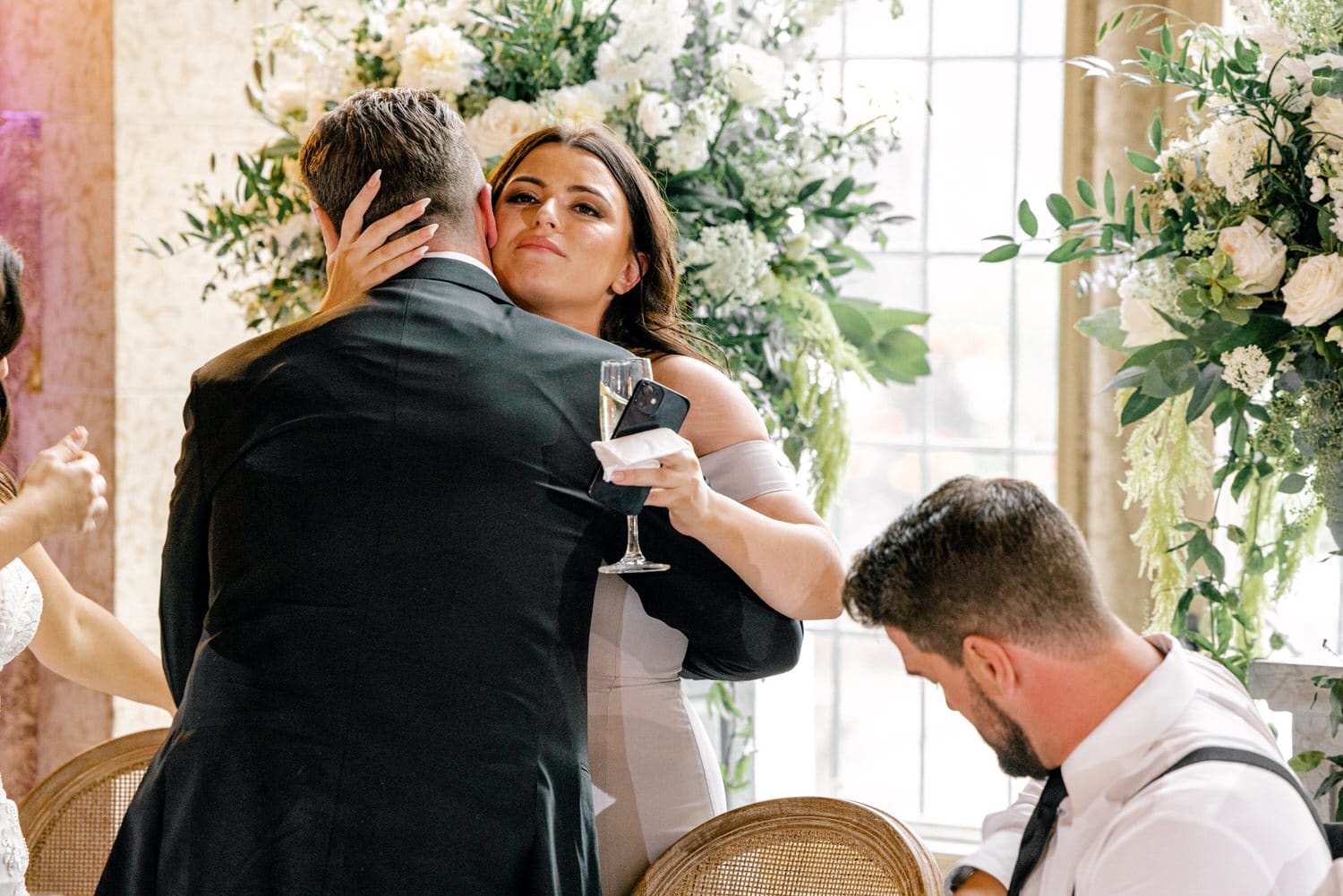 A woman embraces a man during a celebration, holding a glass and a phone, while a man sits nearby, surrounded by floral decorations.