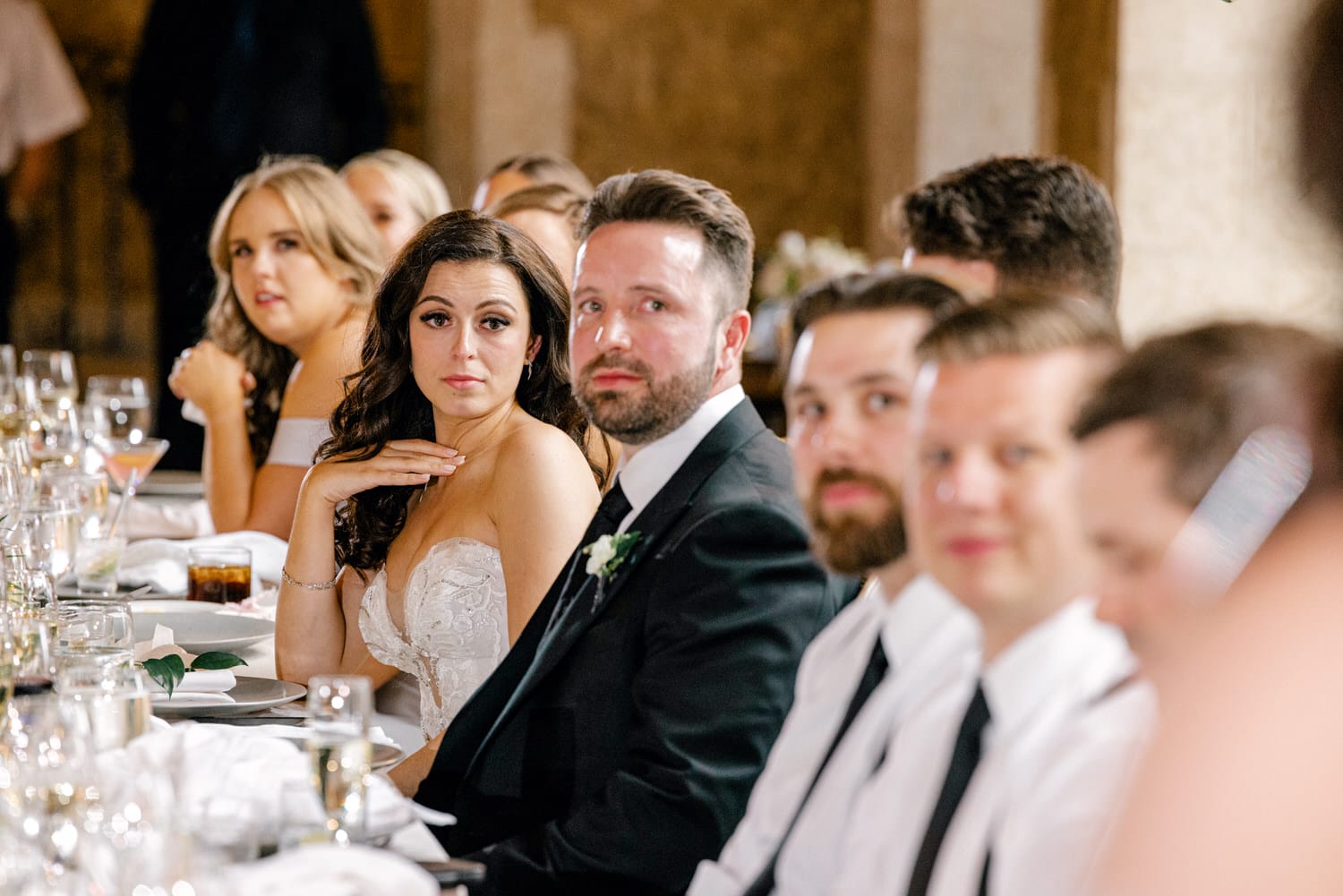 A group of guests seated at a wedding reception, with a bride in a strapless dress and various expressions of engagement and curiosity among the attendees.