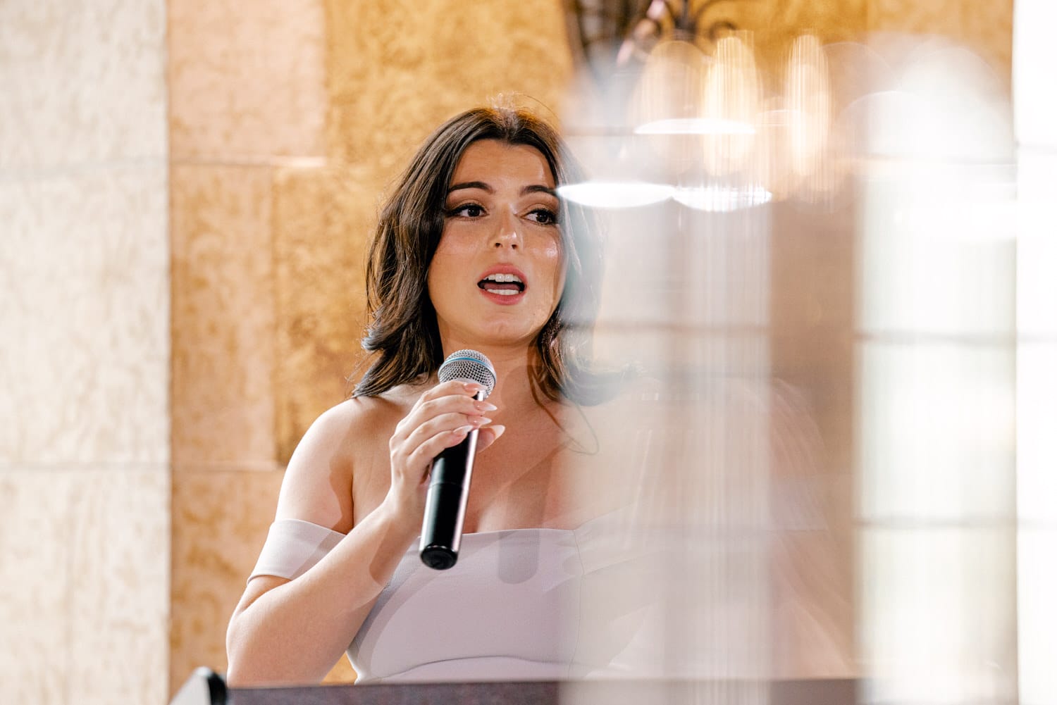 A woman in an elegant off-shoulder dress speaks into a microphone during a wedding reception, captured in a softly blurred setting.