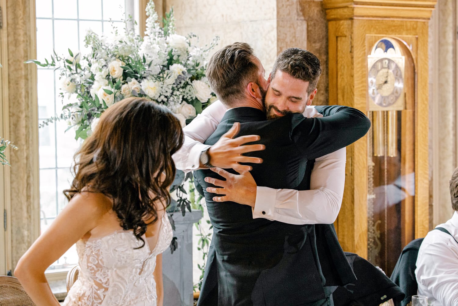 A joyful moment of two men hugging at a wedding, with a bride in the foreground and floral arrangements in the background.