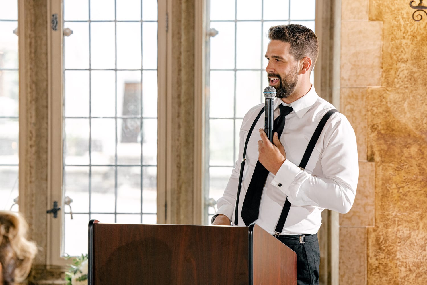 A man in a white shirt and black tie, wearing suspenders, addresses an audience from behind a podium in a well-lit room with large windows.