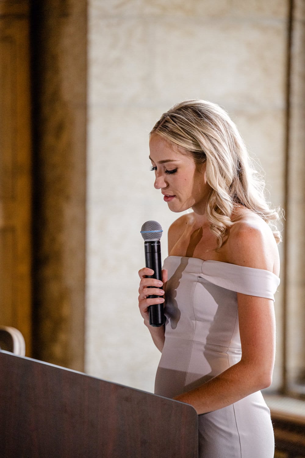 A woman in an elegant dress delivers a heartfelt speech at a formal event, holding a microphone and looking down thoughtfully.