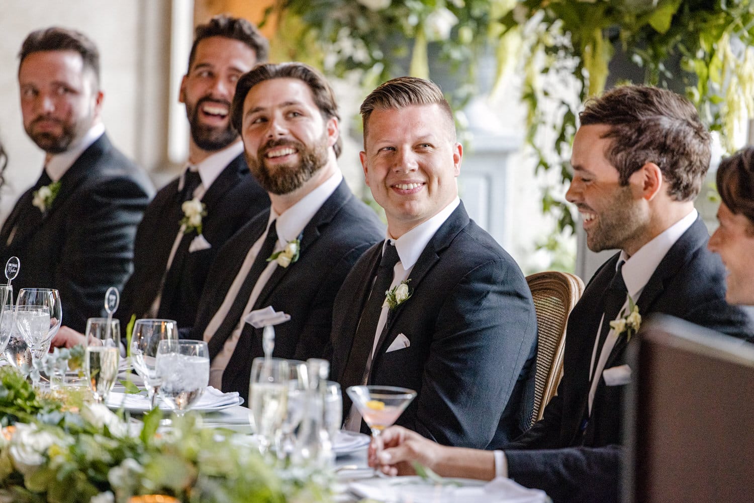 A group of well-dressed men sitting at a elegantly decorated table, smiling and engaging in conversation during a wedding reception.