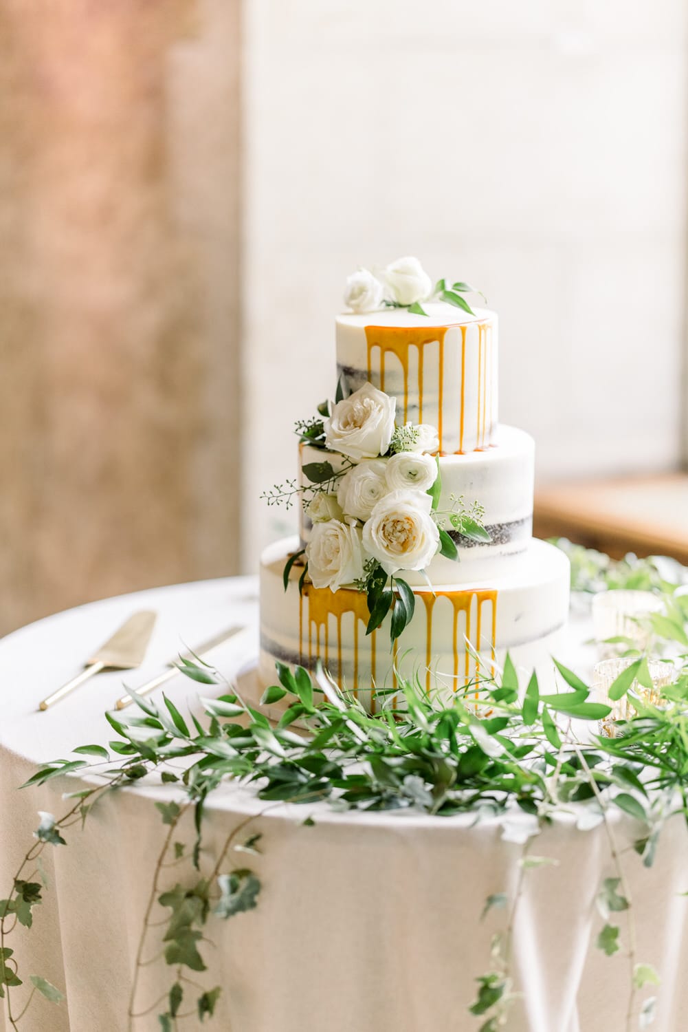 A beautifully designed three-tiered wedding cake adorned with dripping caramel, white roses, and greenery on a table.