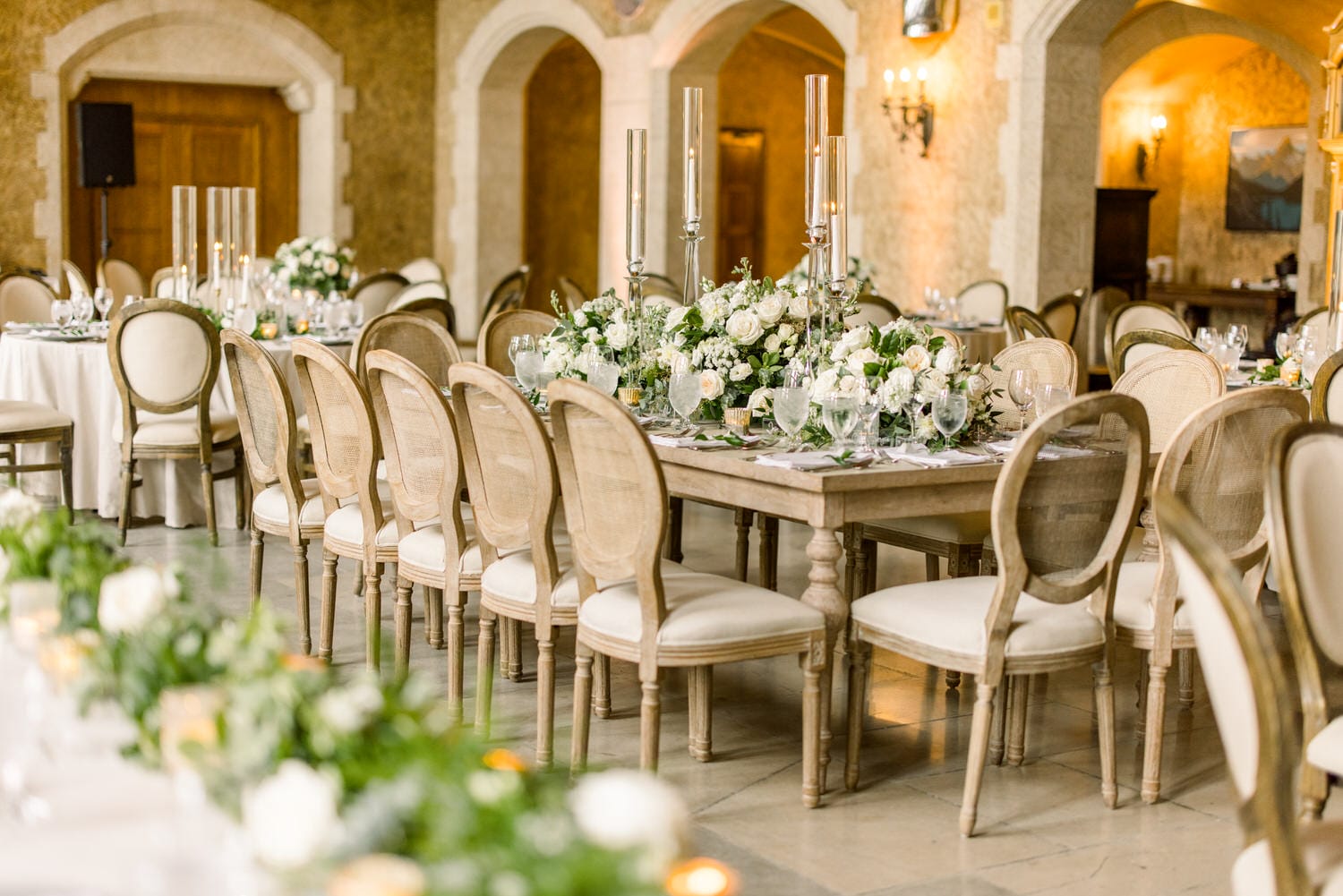 A beautifully arranged dining space featuring wooden tables adorned with floral centerpieces, elegant glassware, and chic chairs in a warmly lit setting.