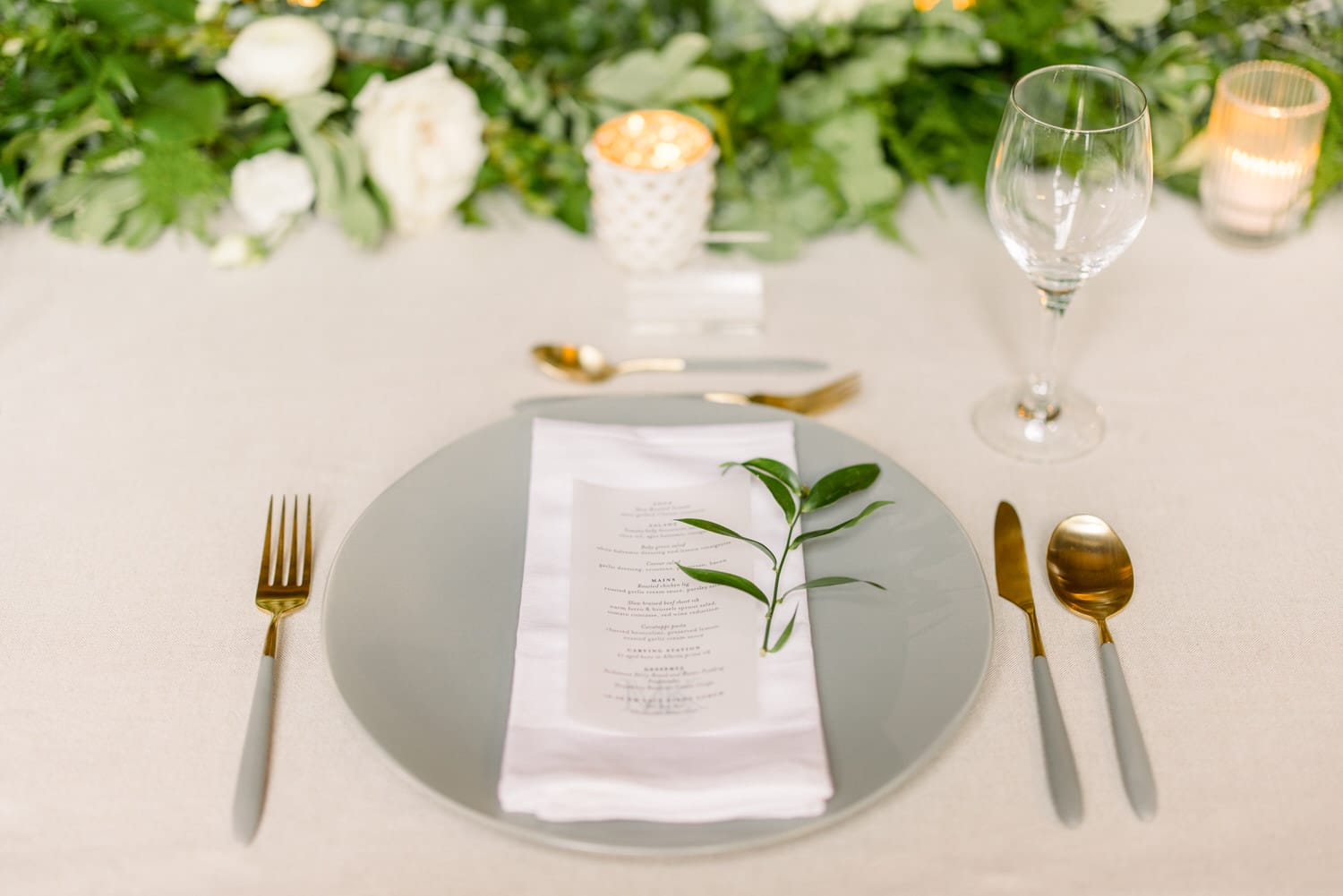 A beautifully arranged dining table featuring a grey plate, gold cutlery, a neatly folded napkin with a menu, and a delicate green branch, surrounded by floral decor and soft candlelight.