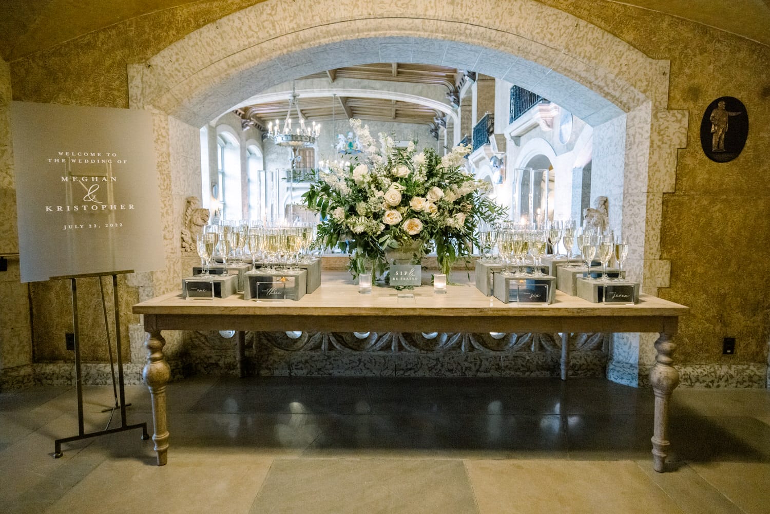 A beautifully arranged table featuring a floral centerpiece, champagne flutes, and a sign welcoming guests to the wedding of Meghan and Kristopher on July 23, 2022.