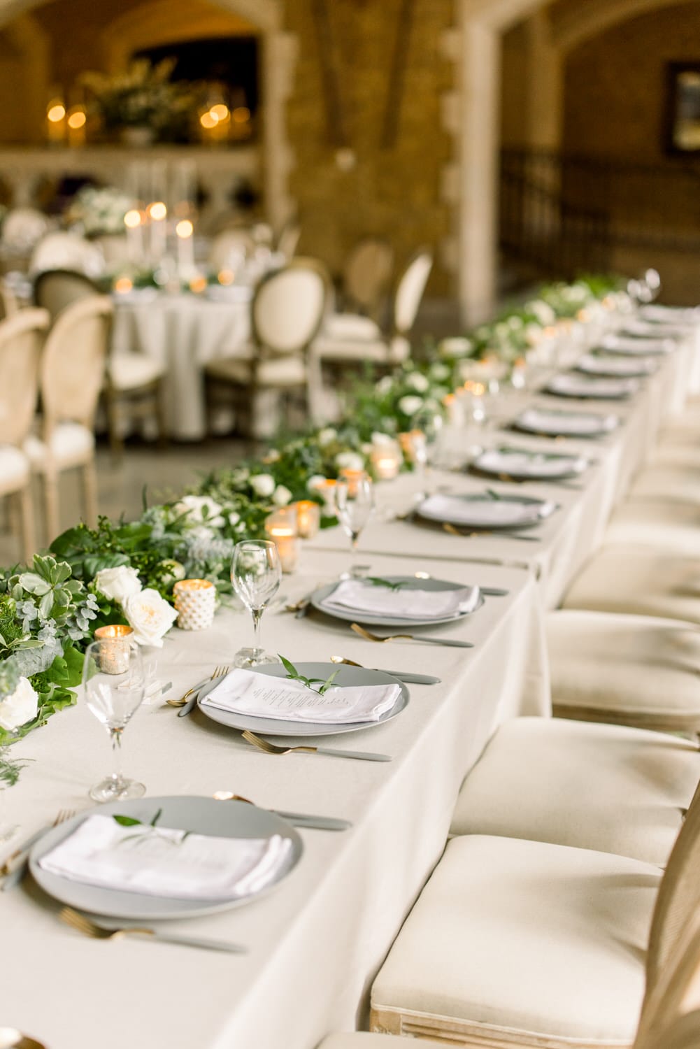 A beautifully arranged dining table featuring gray plates, white napkins, and greenery alongside glowing candles, set in a sophisticated venue.