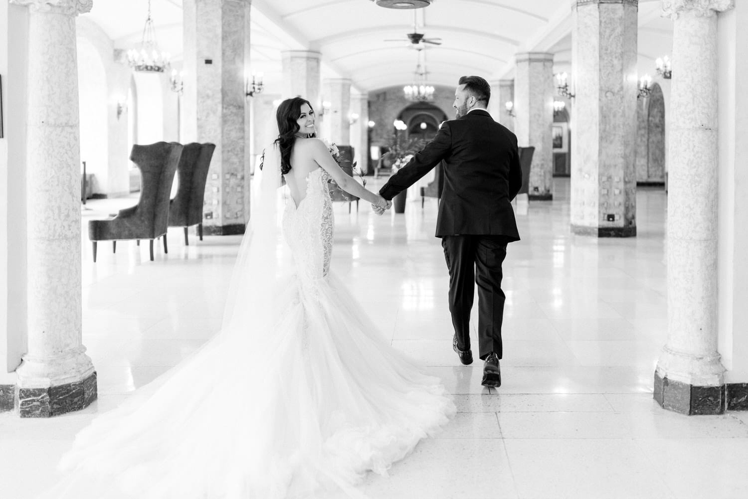 A bride and groom walking hand-in-hand through an elegant, marble-floored hallway, capturing a moment of joy and love in black and white.