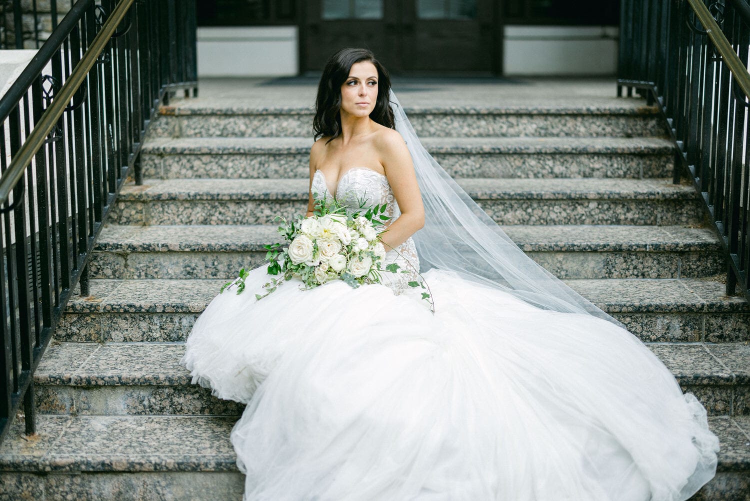 A bride in a stunning white gown and veil sits gracefully on stone steps, holding a bouquet of white flowers and greenery.