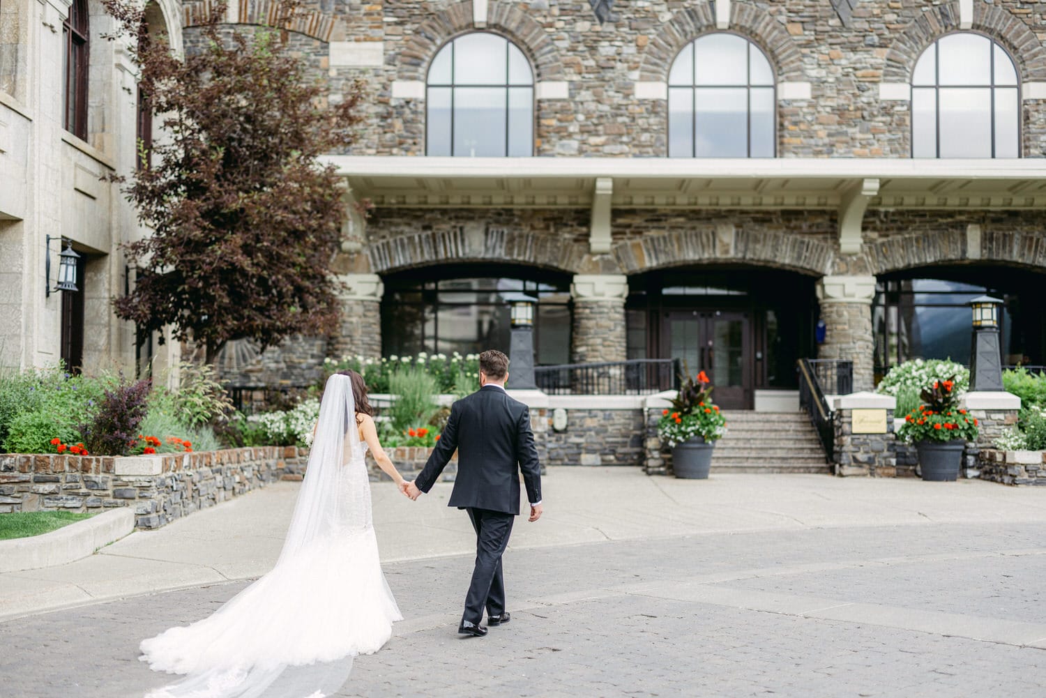 A bride and groom walk hand in hand toward a stone building adorned with greenery and colorful flowers.