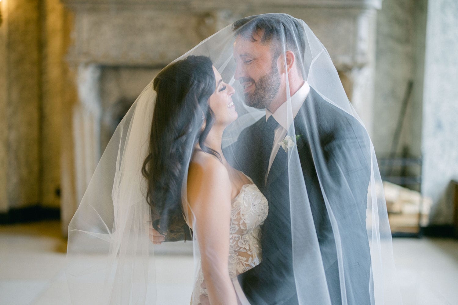 A couple sharing a joyful moment together, faces close, enveloped in a soft wedding veil, in an elegant indoor setting.