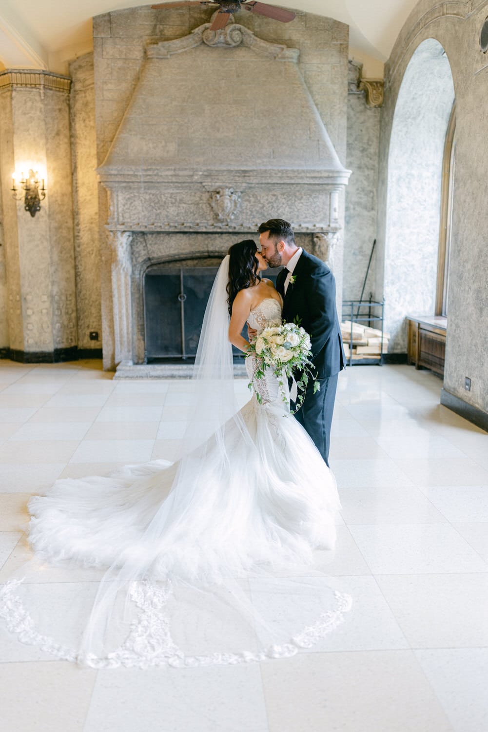 A bride and groom share a tender kiss in an elegant indoor setting, framed by an ornate fireplace and large windows.