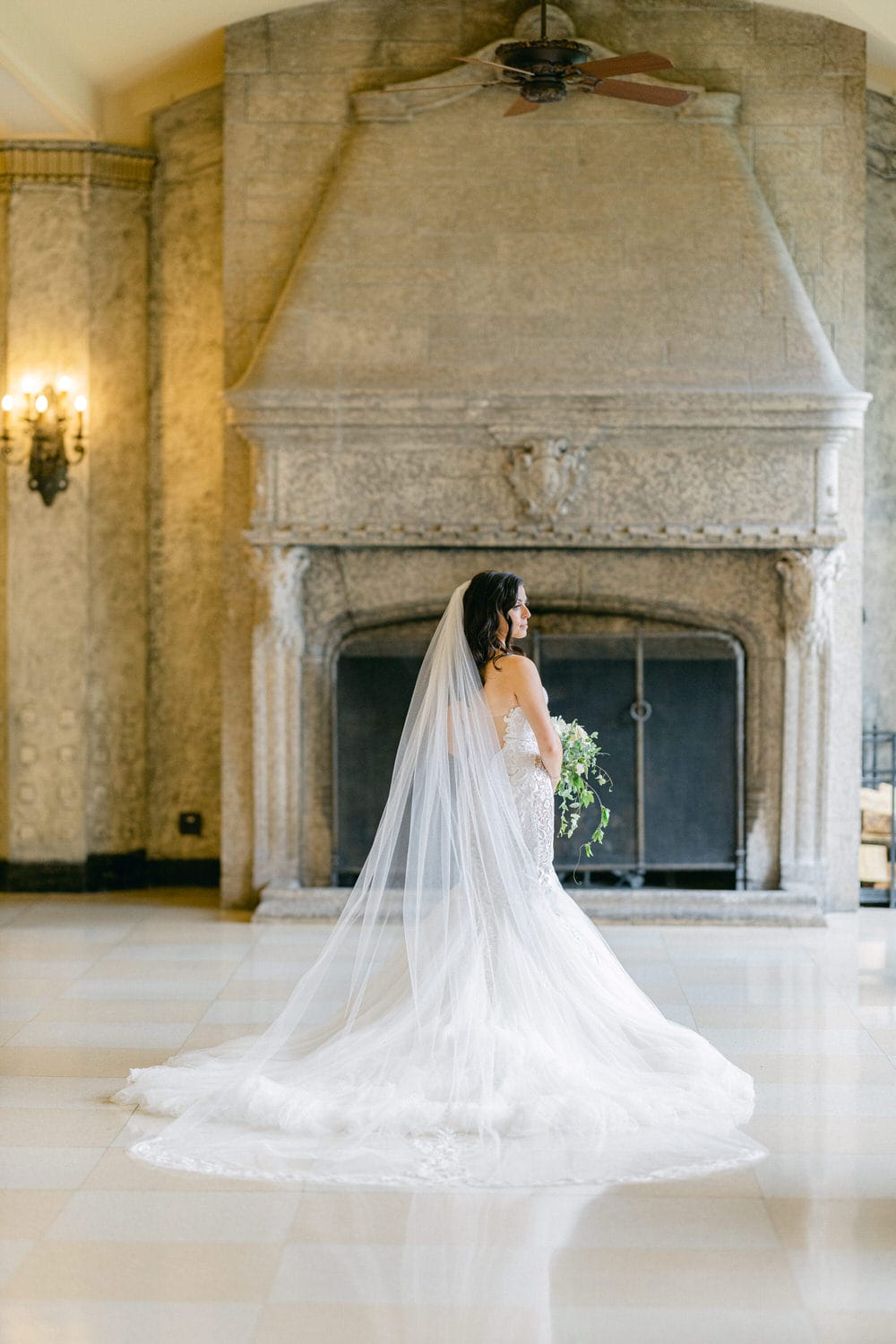 A bride in a stunning wedding dress stands gracefully in a historic room, showcasing her elegant veil and bouquet, with a grand fireplace in the background.