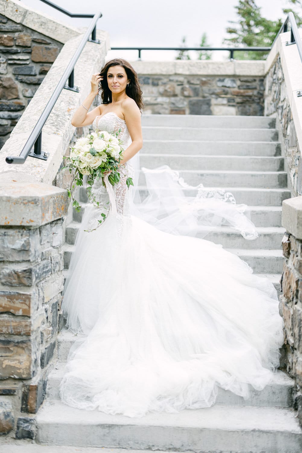 A beautiful bride in a stunning white wedding gown poses gracefully on stone steps, holding a bouquet of white flowers, with a flowing veil accentuating her look.