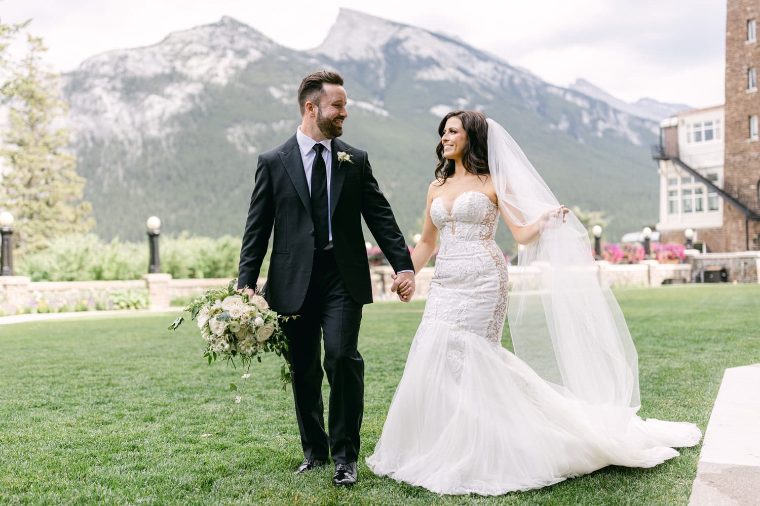 A joyful couple walks hand-in-hand on a grassy lawn, surrounded by mountains, with the bride in a beautiful lace wedding gown and the groom in a classic tuxedo, holding a bouquet of white roses.