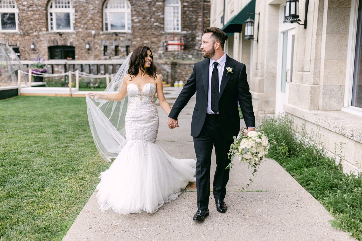 A bride in an elegant gown and a groom in a suit walk hand in hand, smiling at each other in a picturesque outdoor setting.