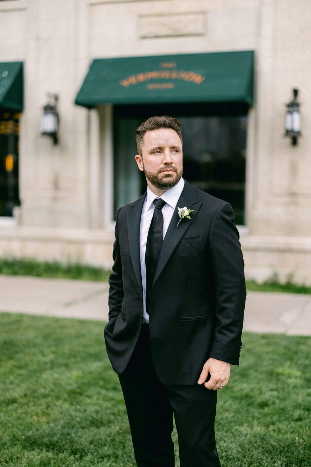 A man in a black tuxedo stands confidently outdoors, wearing a white dress shirt and a small flower boutonniere, with a blurred building in the background.