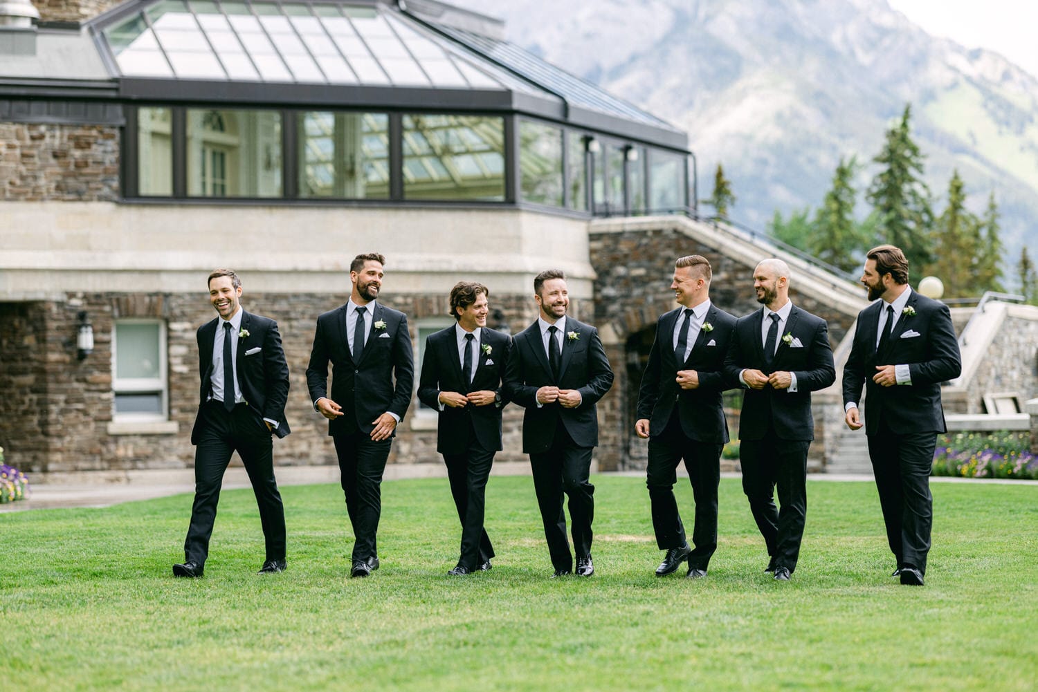 A group of seven well-dressed groomsmen in black suits and ties, joyfully walking on a grassy lawn in front of a stone building surrounded by trees and mountains.