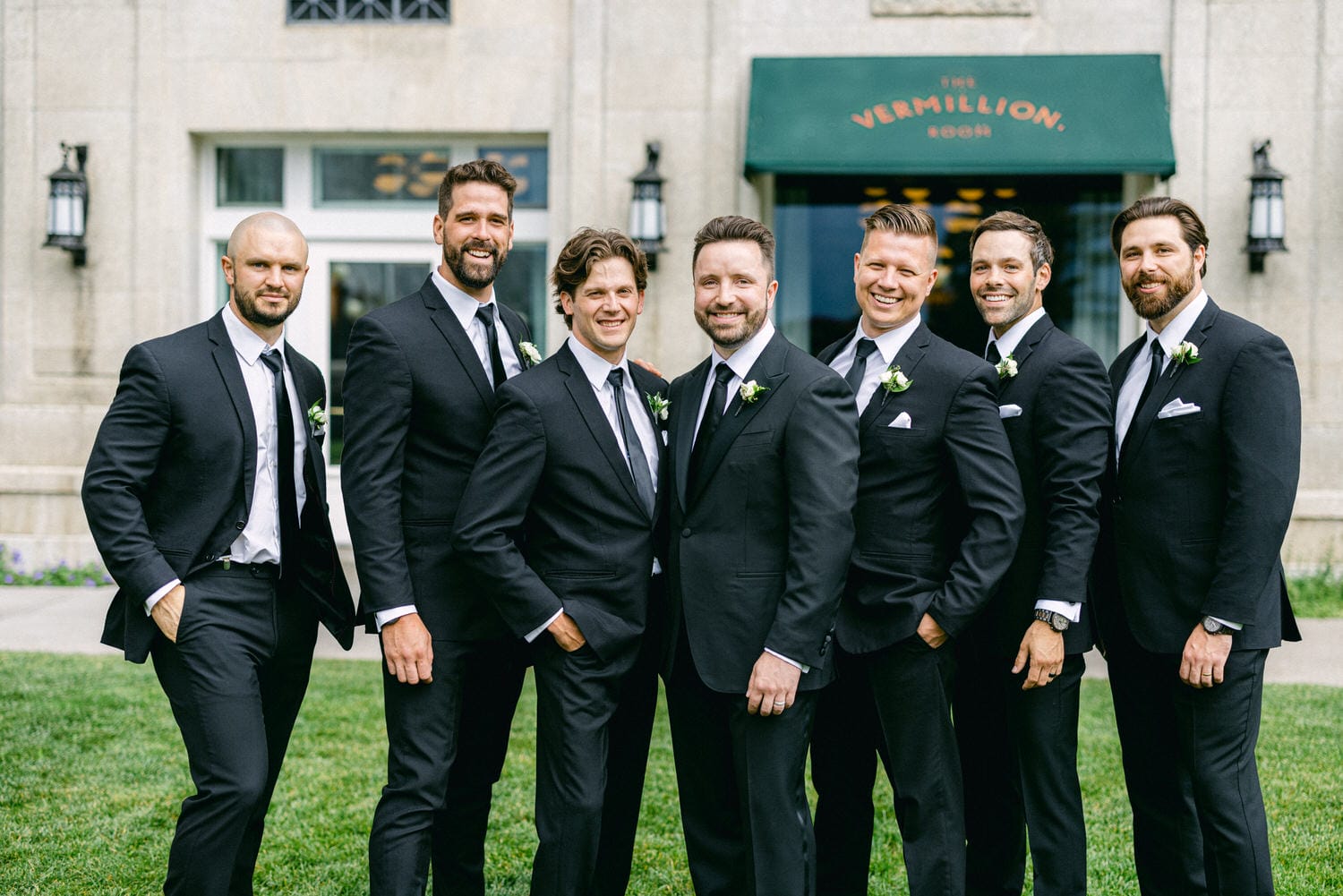 A group of seven well-dressed men in black suits with white flowers, posing confidently outdoors in front of a building with a green awning.