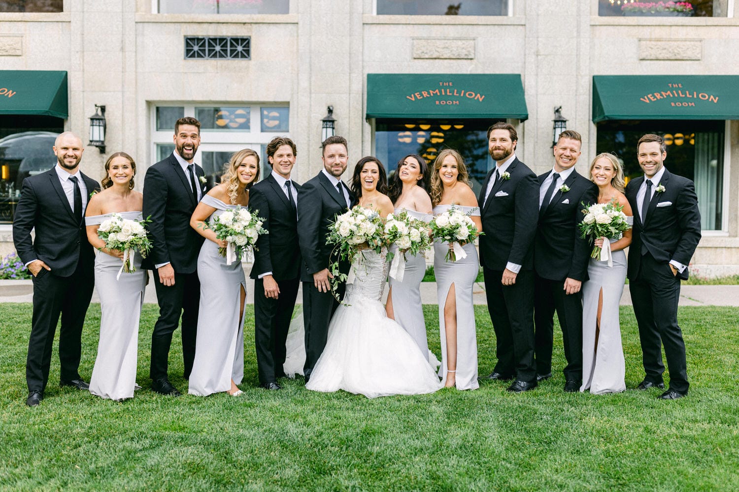 A cheerful wedding party poses together outside the Vermillion Room, with the bride in a stunning gown and the bridal party in elegant light-gray dresses, all holding lush floral bouquets.