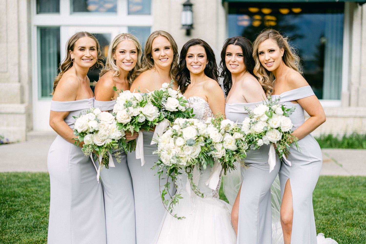 A joyous bridal party poses together, showcasing elegant off-shoulder grey dresses and lush white floral bouquets against a picturesque venue backdrop.