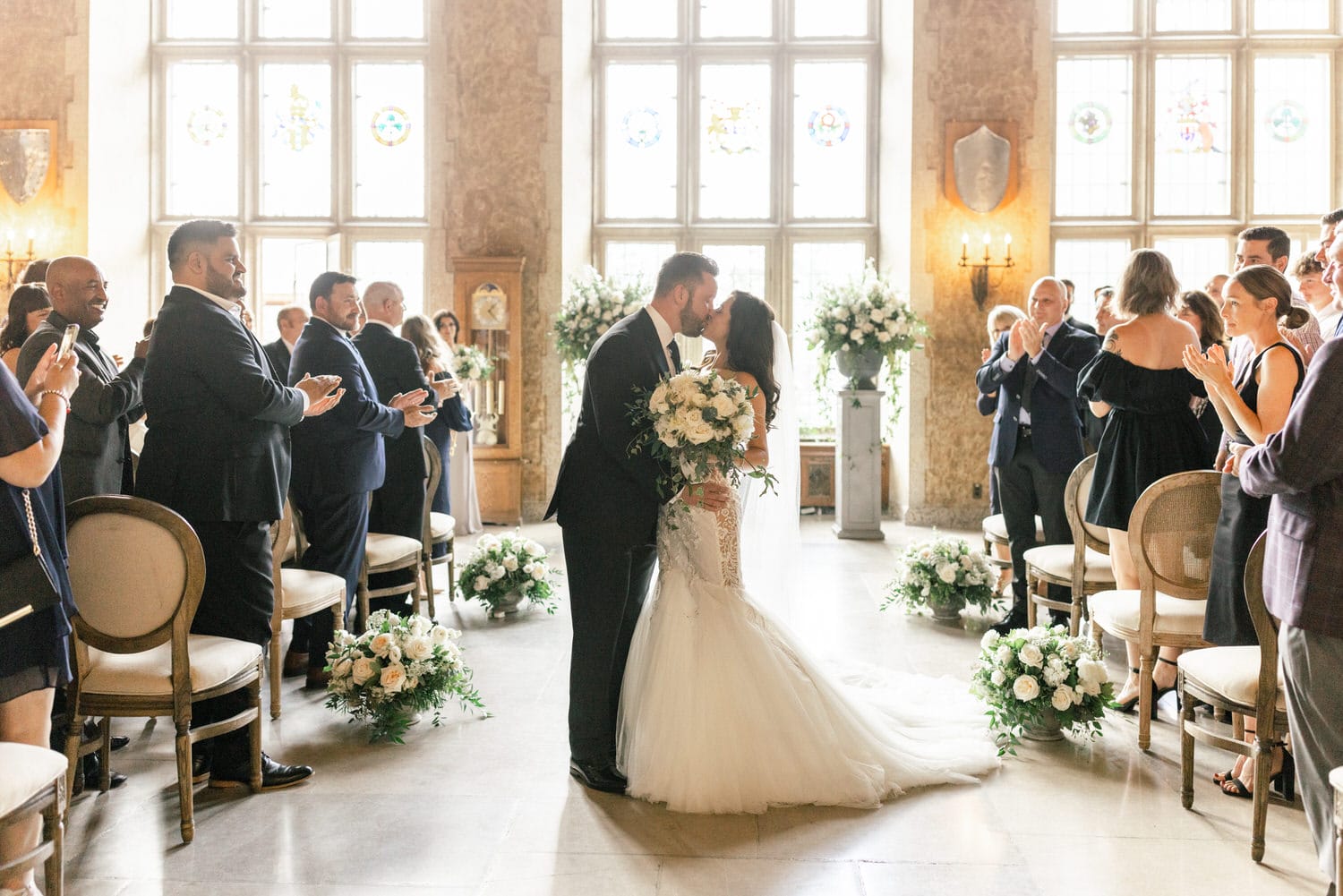 A couple shares a kiss during their wedding ceremony, surrounded by guests applauding in a beautifully decorated hall with floral arrangements and stained-glass windows.