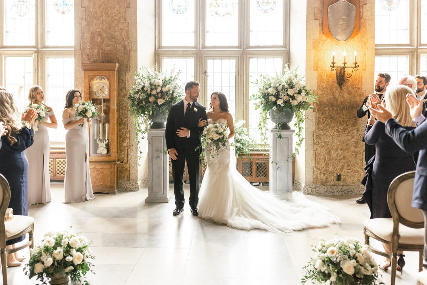 A couple, adorned in elegant attire, smiles at each other during their wedding ceremony, surrounded by floral arrangements and guests applauding.