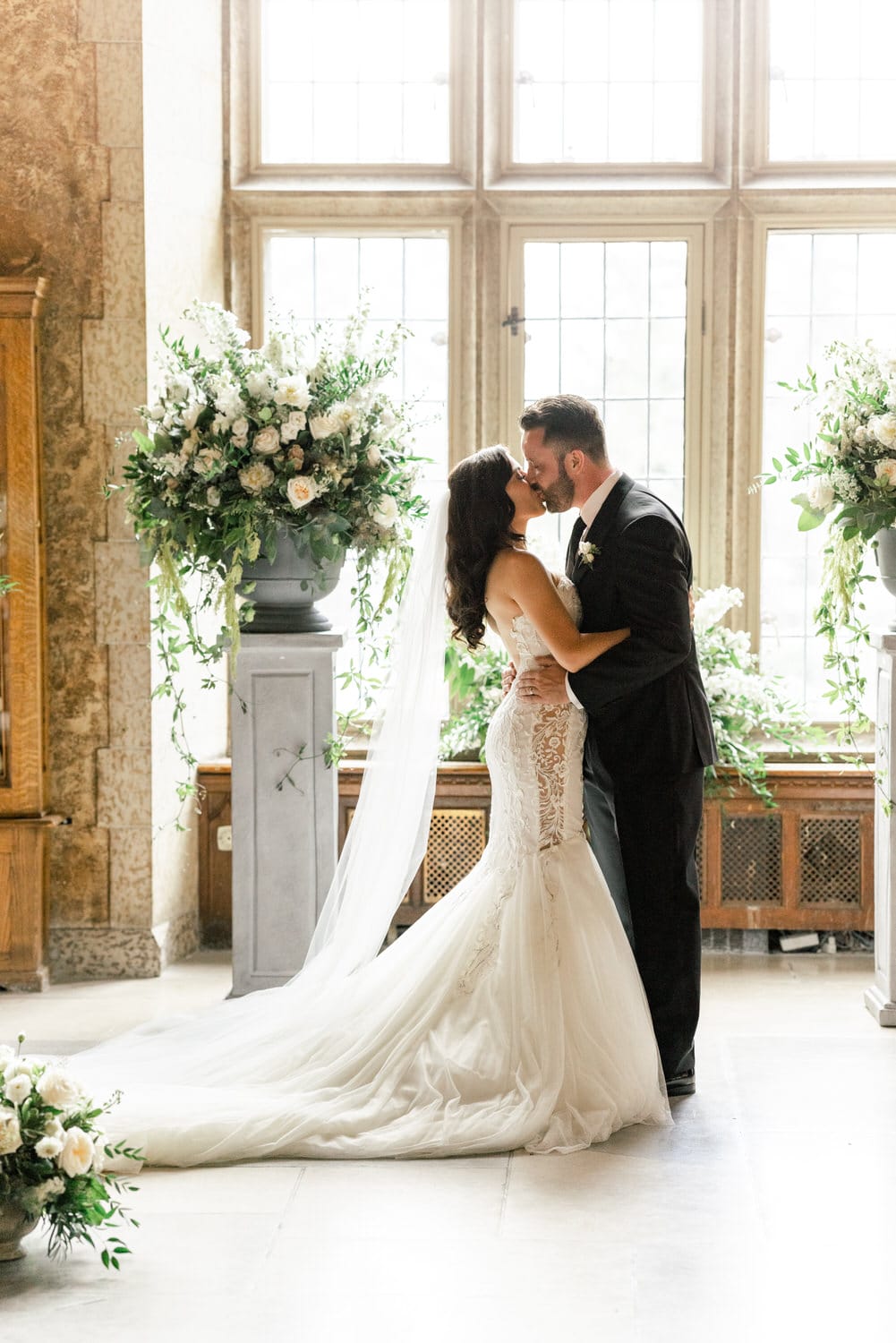 A bride and groom share a tender kiss in a beautifully decorated space, surrounded by floral arrangements and soft natural light.