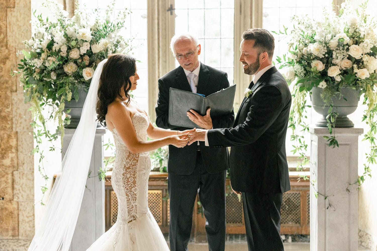 A bride and groom exchanging rings during their wedding ceremony, with a celebrant officiating in an elegant setting adorned with floral arrangements.