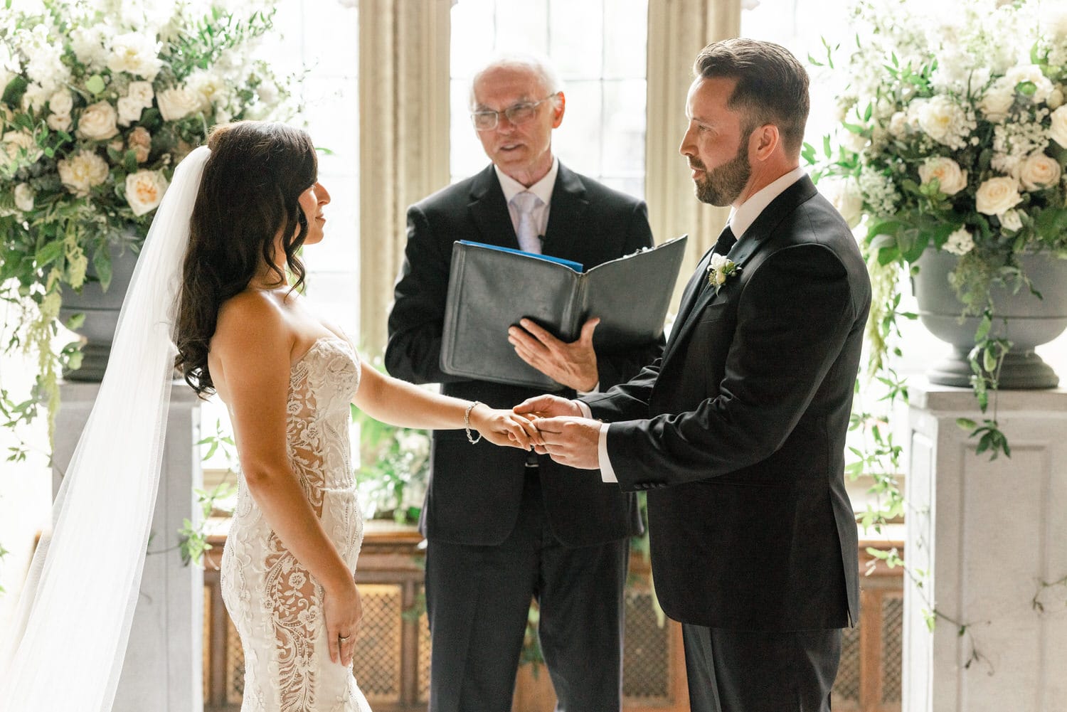 A bride and groom exchange vows during their wedding ceremony, with an officiant in the background amidst a floral arrangement.