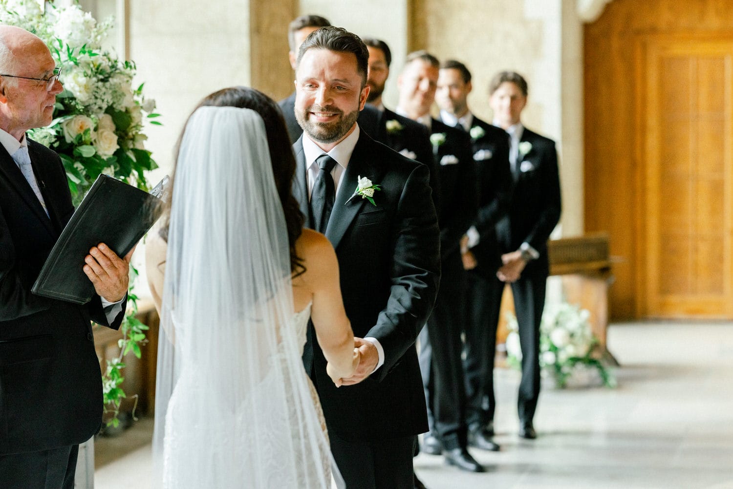 A couple exchanging vows during a wedding ceremony, with the officiant and groomsmen in the background.