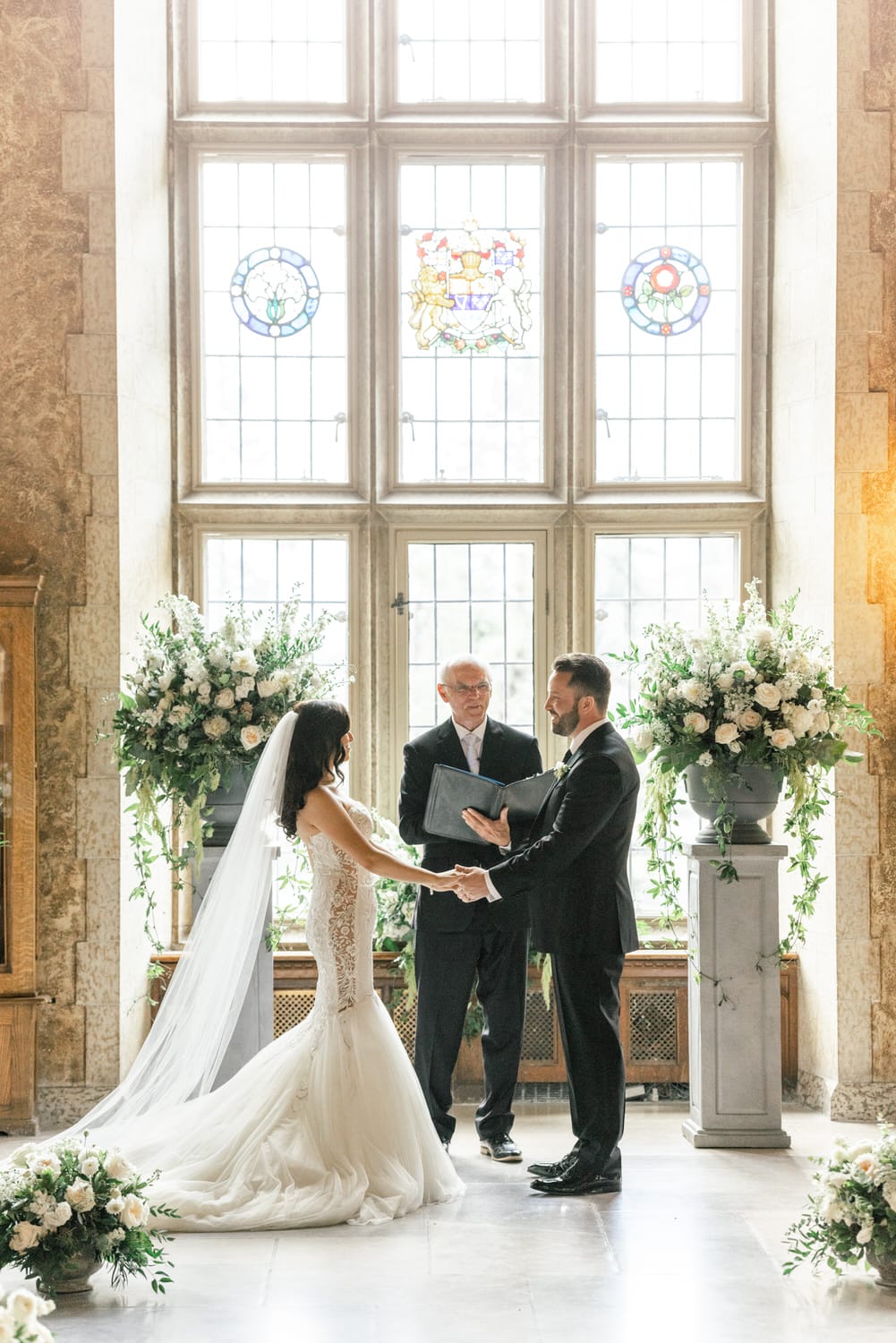 A bride and groom exchange vows in a beautifully decorated historic venue, featuring stained glass windows and floral arrangements.