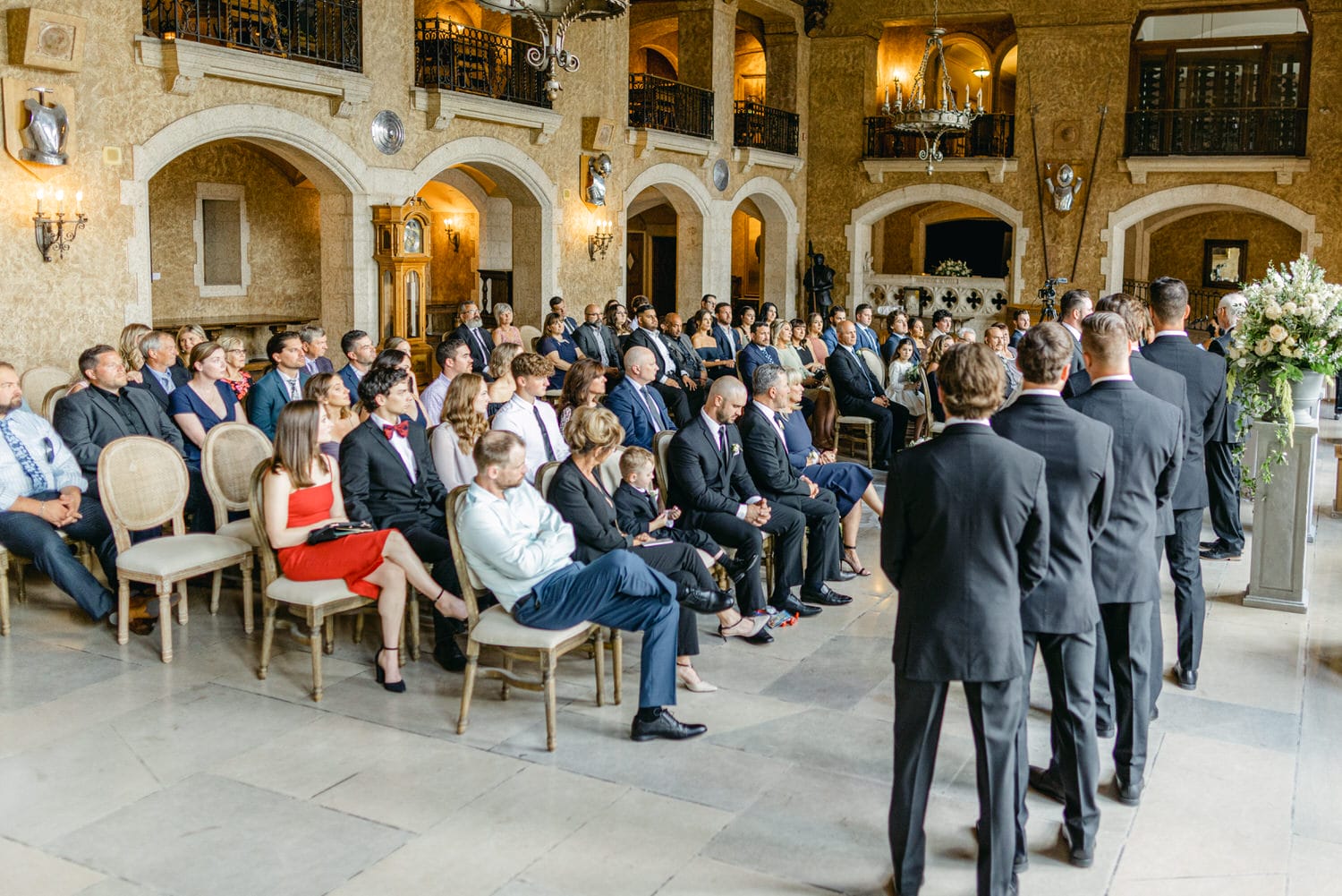 A diverse group of guests seated during a formal wedding ceremony in a beautifully decorated hall, with a focus on the attendees and the wedding party standing at the front.