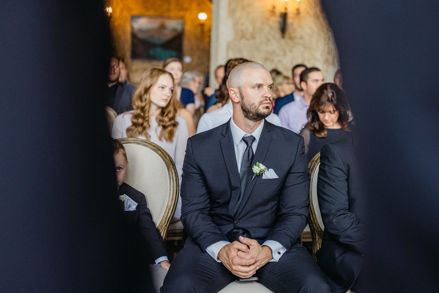 A groom seated thoughtfully among guests at a wedding ceremony, showcasing a moment of anticipation and emotion.