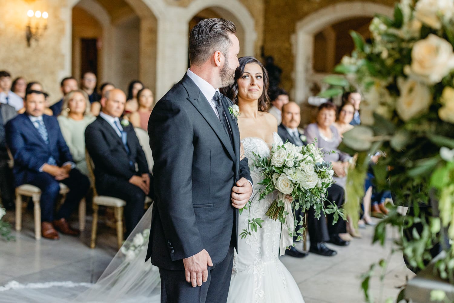 Bride at the altar smiling at her groom during a wedding ceremony, surrounded by guests and floral arrangements.