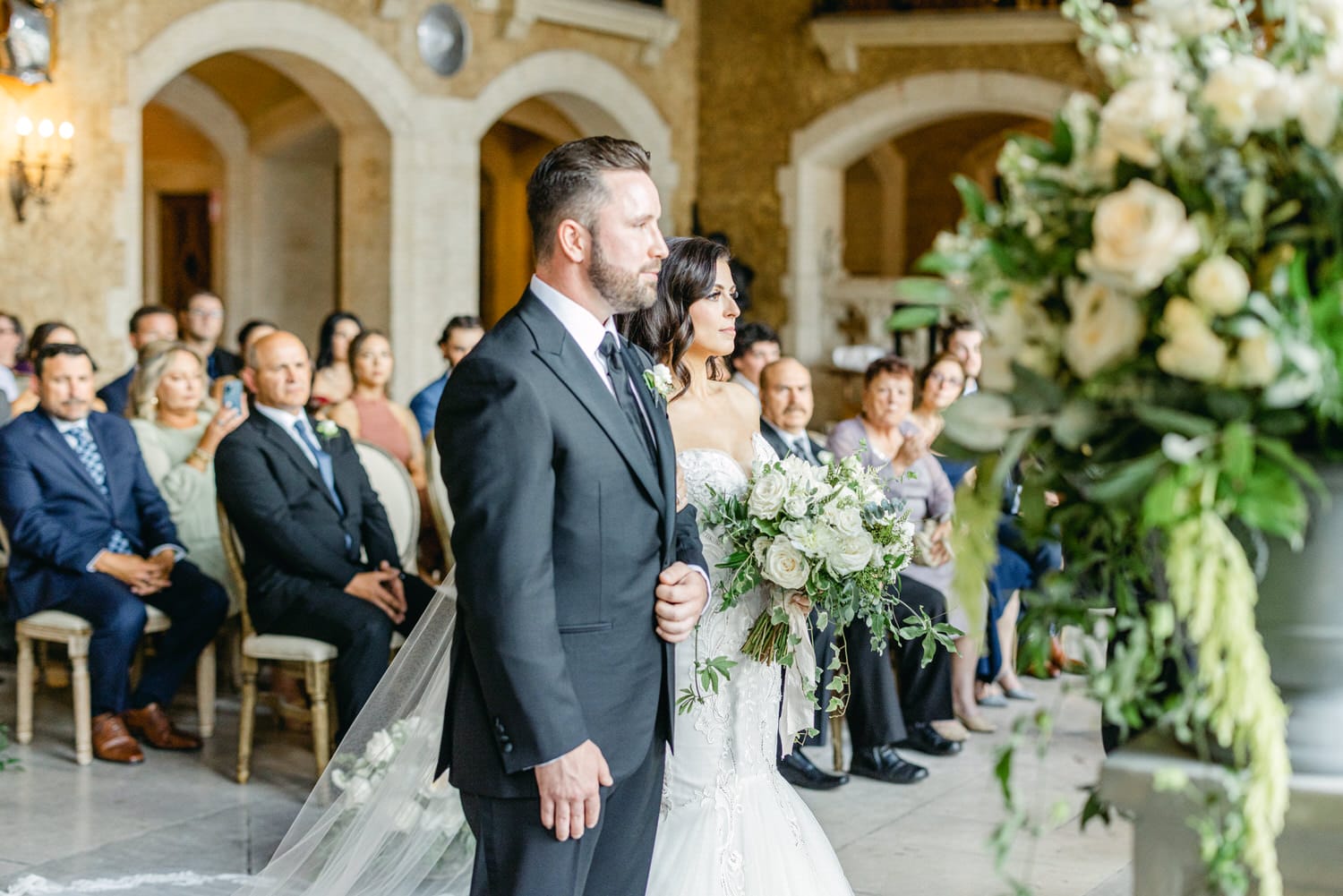 A bride and groom stand together during their wedding ceremony, surrounded by guests in an elegantly decorated venue. The bride holds a bouquet of white roses and greenery, while the groom stands beside her, exuding a sense of joy and commitment.