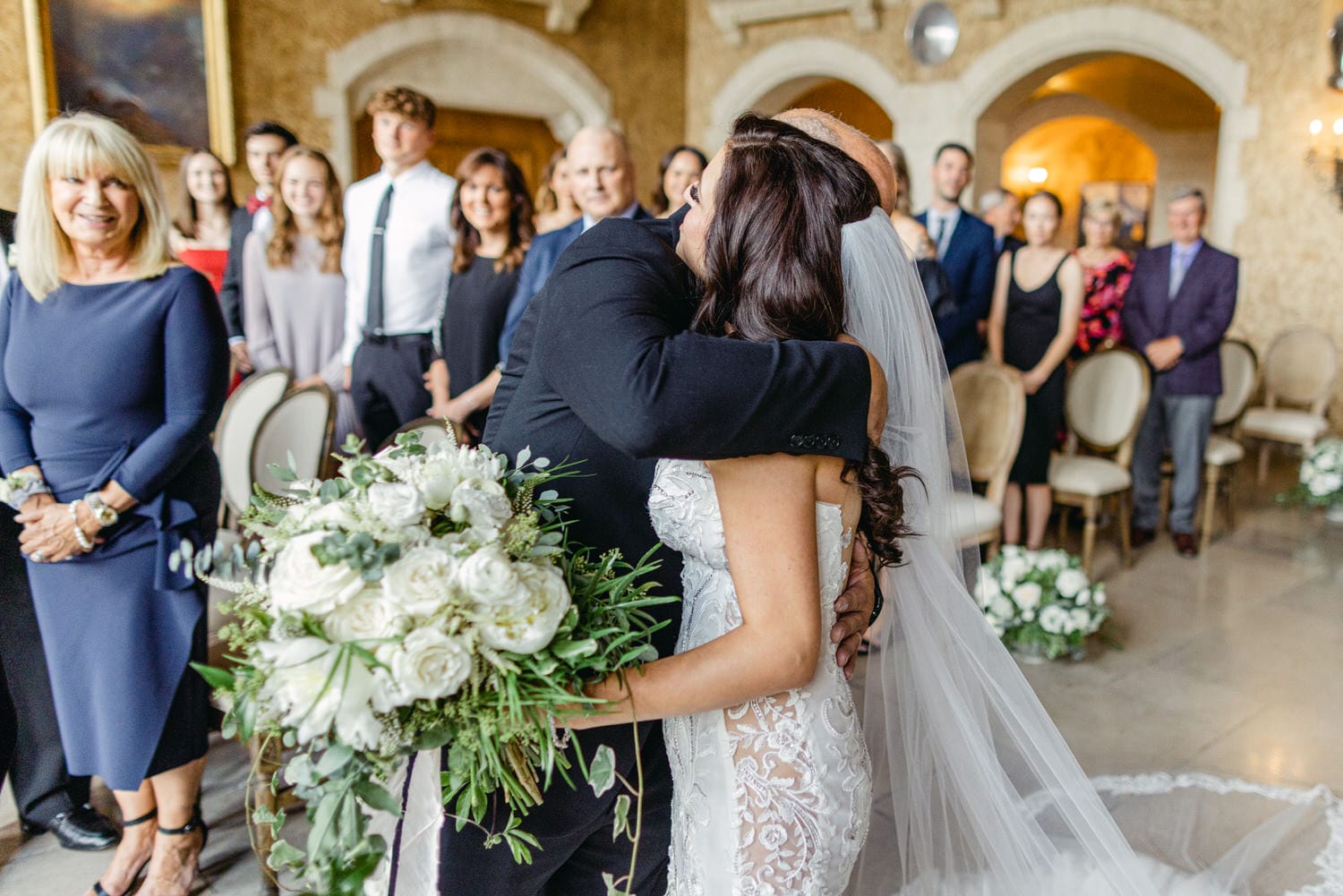 A bride embraces a loved one during a wedding ceremony, surrounded by guests and floral arrangements.