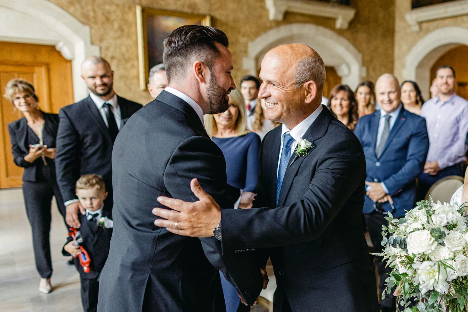 A groom joyfully greets a smiling father figure during a wedding ceremony, surrounded by guests in elegant attire.