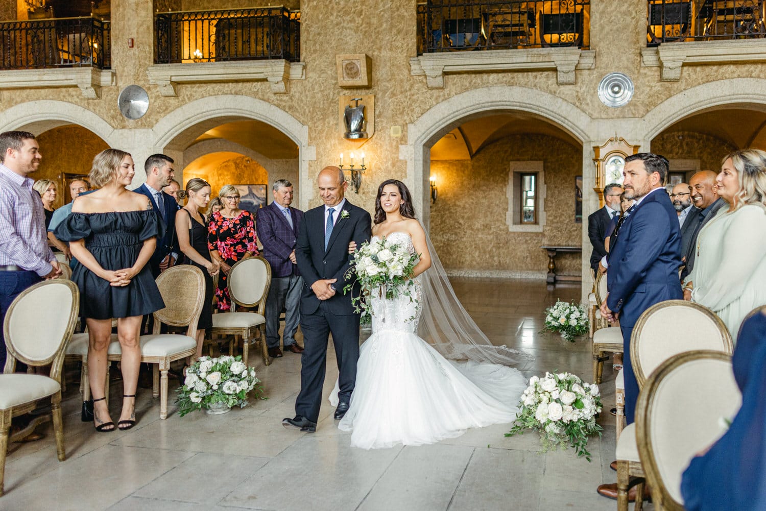 A bride walks down the aisle with her father, surrounded by guests in an elegant venue, highlighting a moment of love and celebration.