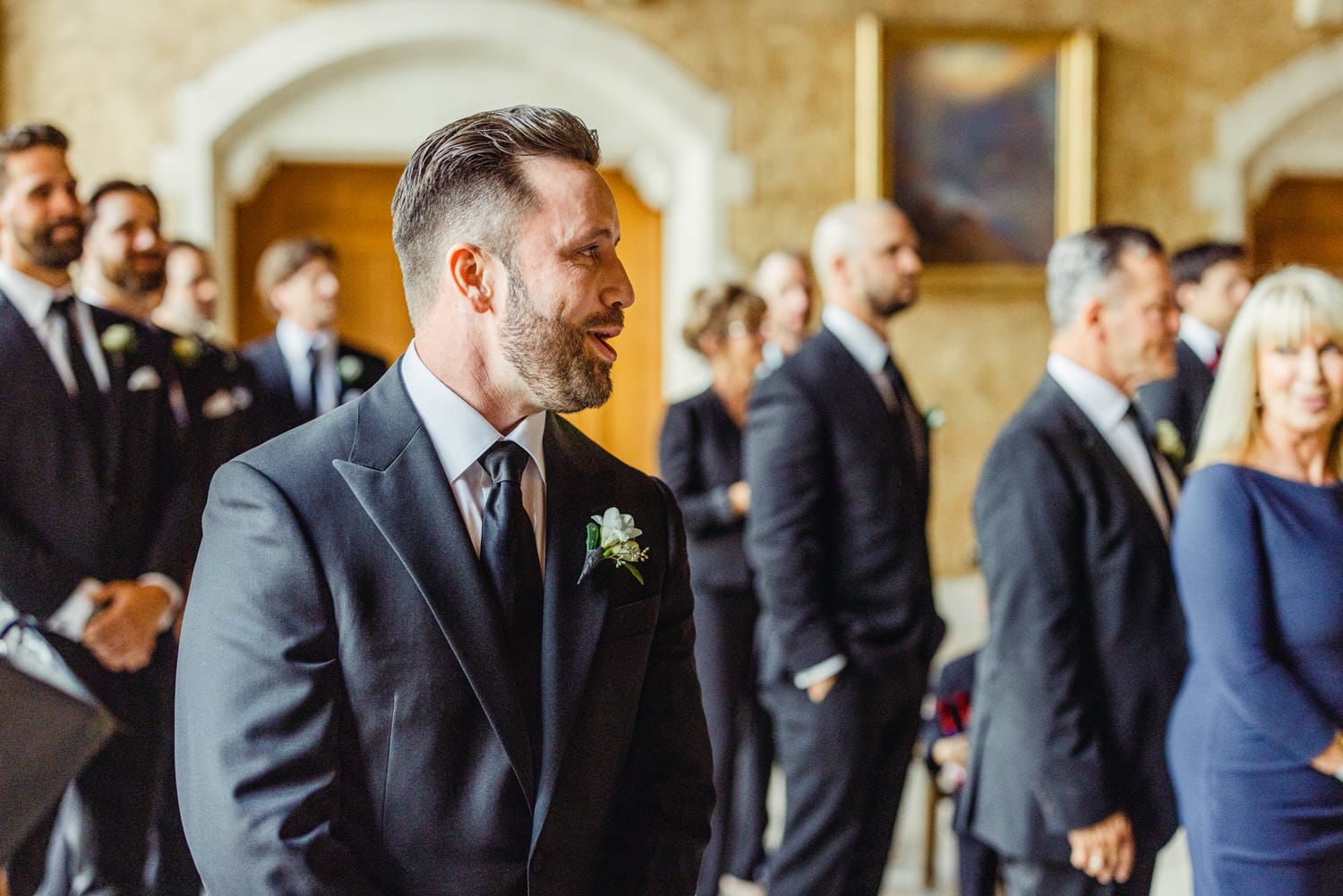 A man in a tuxedo smiles joyfully during a wedding ceremony, surrounded by well-dressed guests.