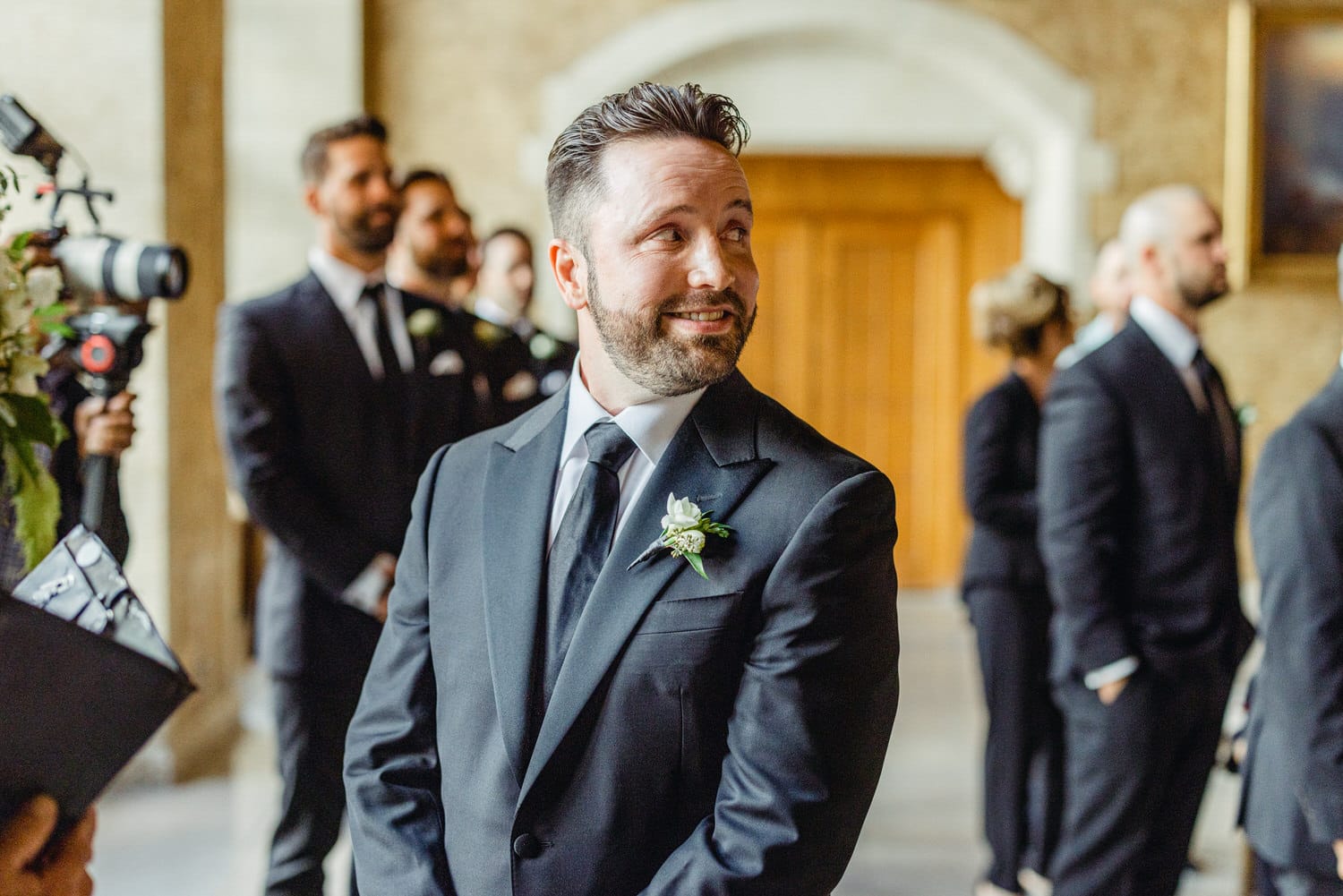 A groom in a sharp black suit, smiling and looking back with joy in a festive setting, surrounded by groomsmen in formal attire.