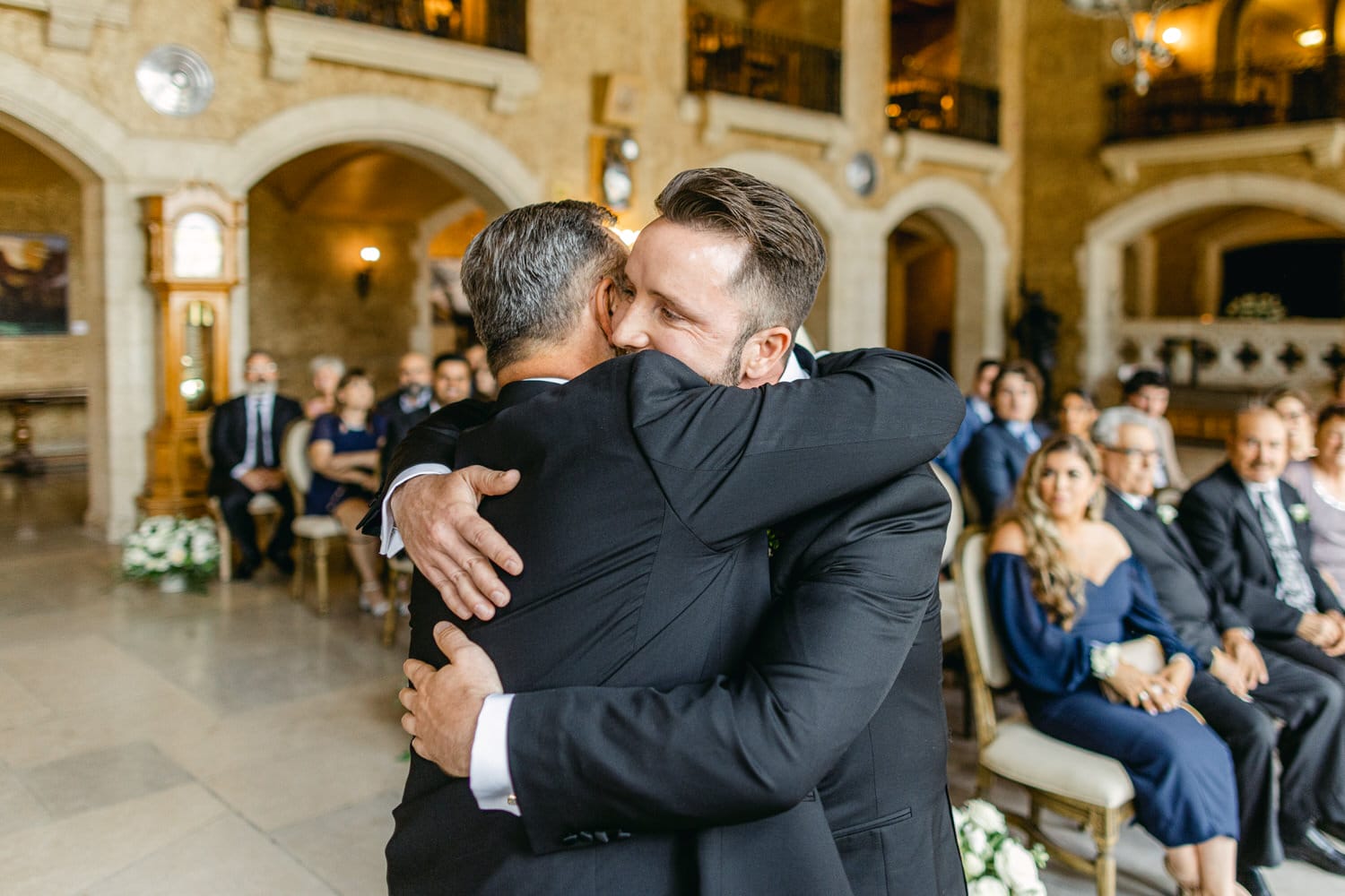 Two men share a heartfelt hug during a wedding ceremony, surrounded by seated guests in a beautifully decorated venue.