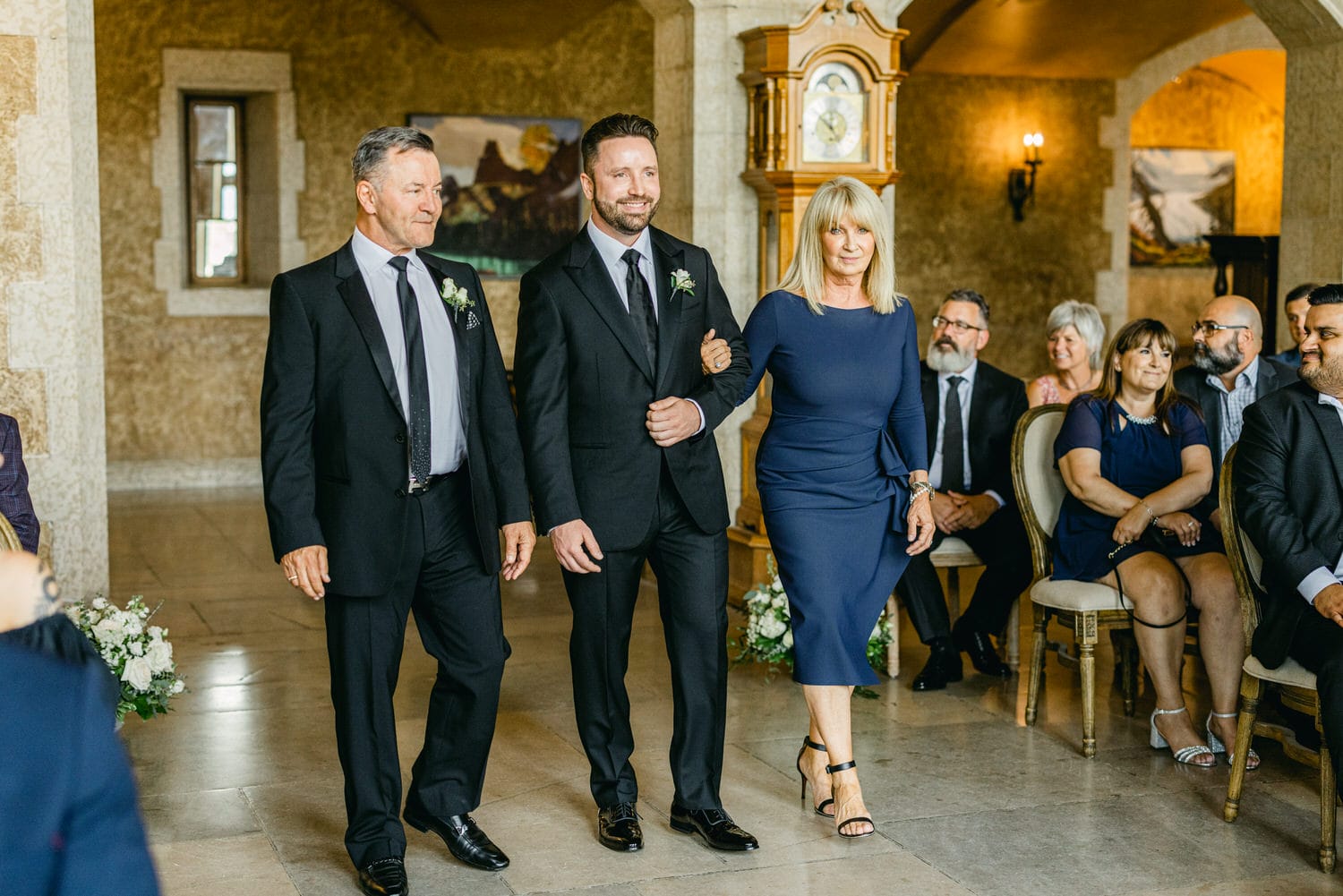 A groom walking down the aisle with his parents, surrounded by guests in a beautifully decorated venue.