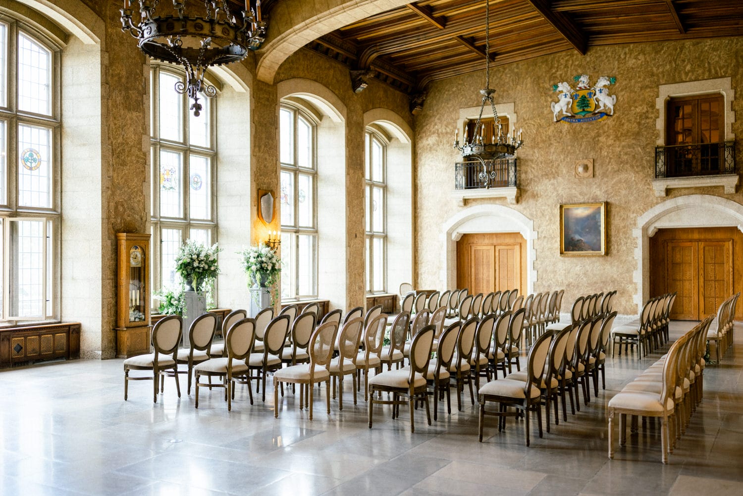 A spacious, beautifully decorated room featuring rows of chairs, large windows, floral arrangements, and ornate chandeliers, set for an event.