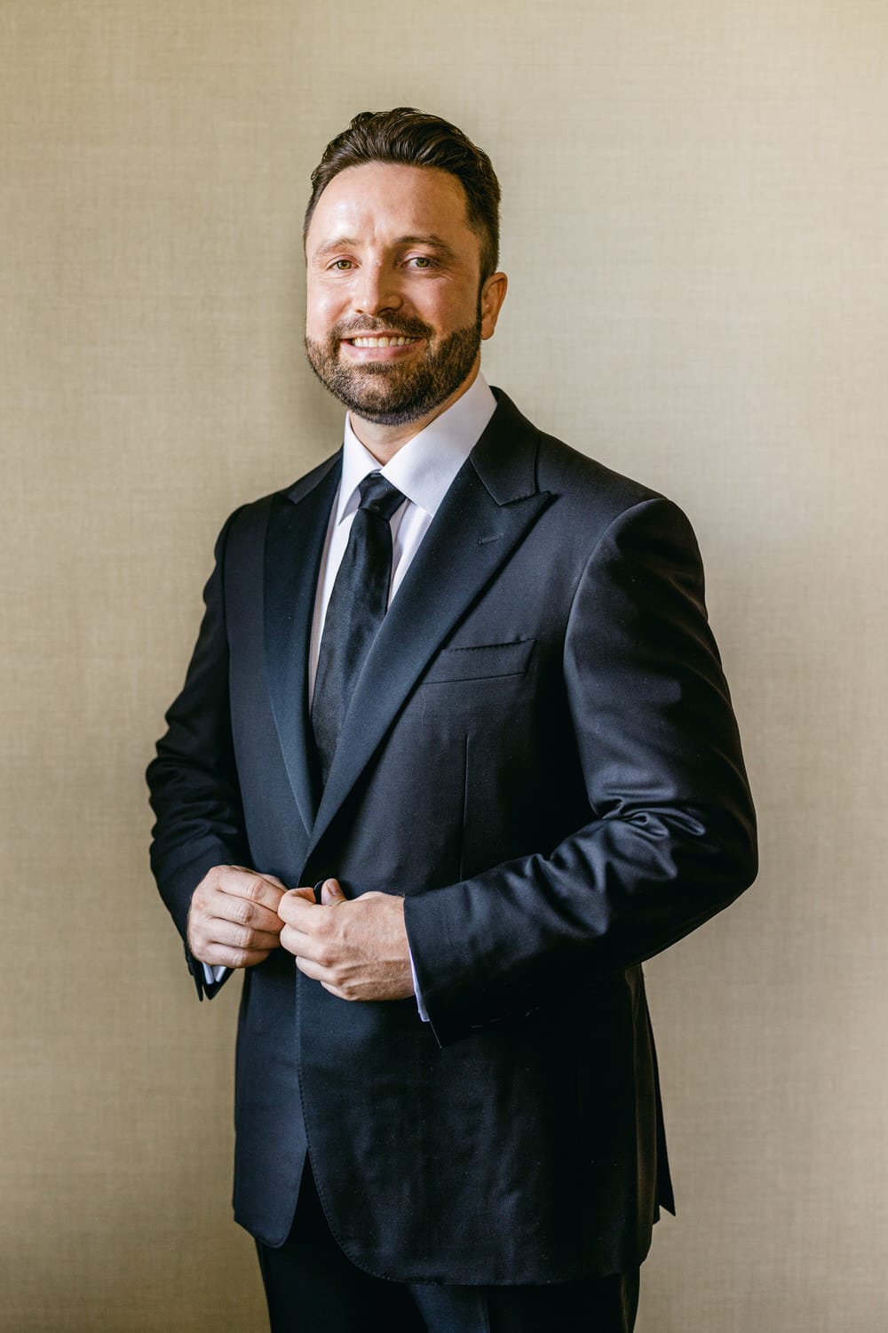 A smiling man in a black suit adjusting his jacket, standing in front of a neutral textured background.