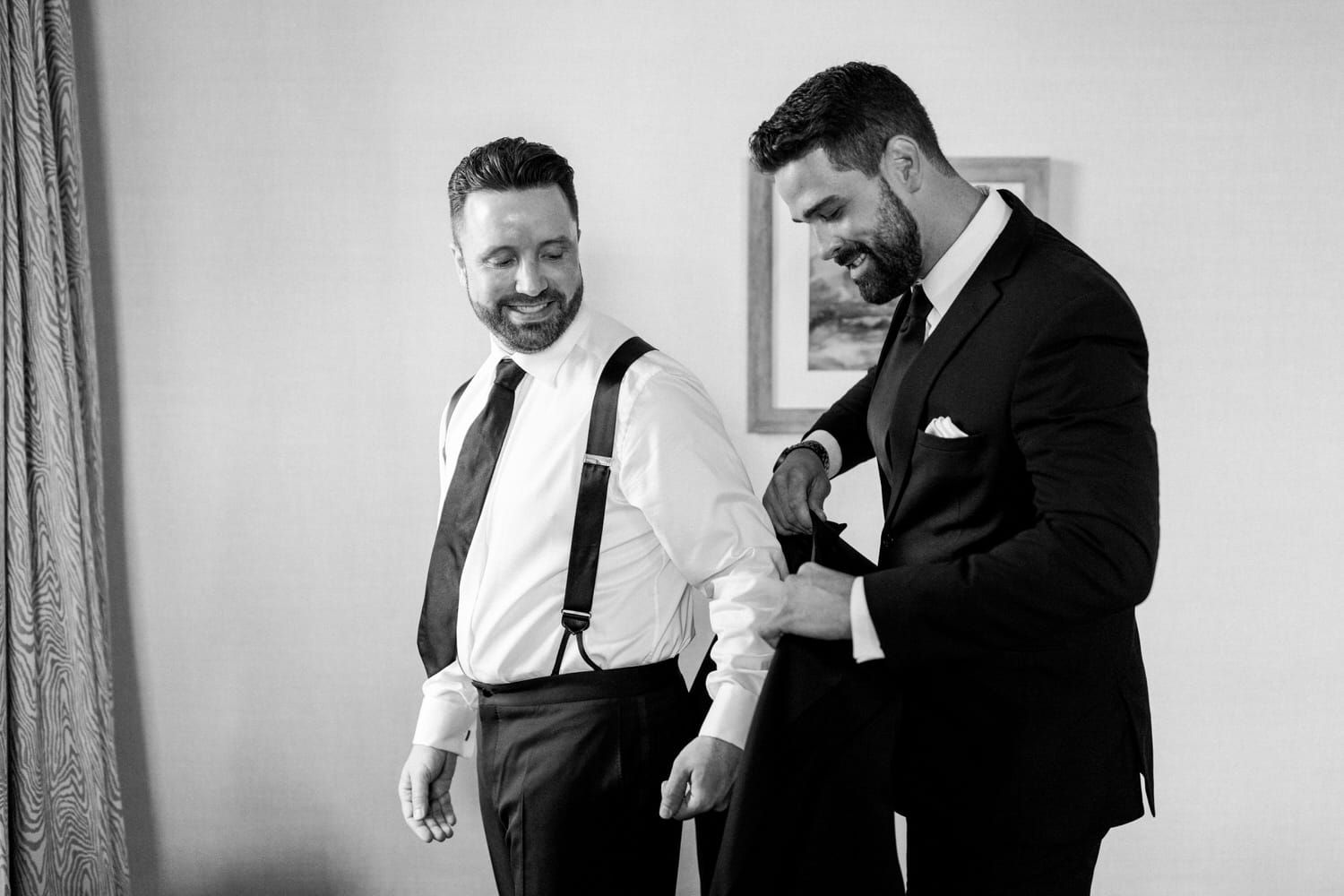 Two men assist each other getting ready in formal attire, showcasing smiles and camaraderie in a well-lit room.