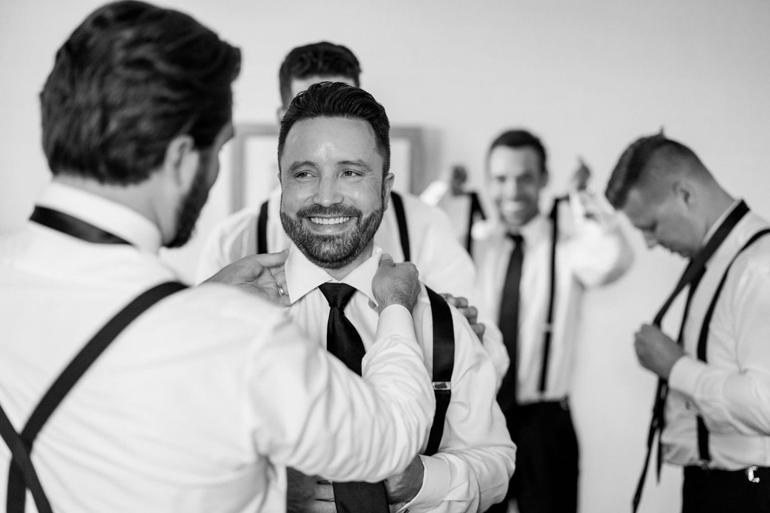 A joyful groom getting ready with the help of his groomsmen, all dressed in white shirts and black ties, capturing a moment of camaraderie and excitement.